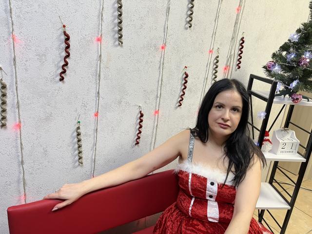 TayaBoret with black hair, red dress with white fur trim and red button, sitting on red bench, holding bench, Christmas tree and decorations in background, smiling.