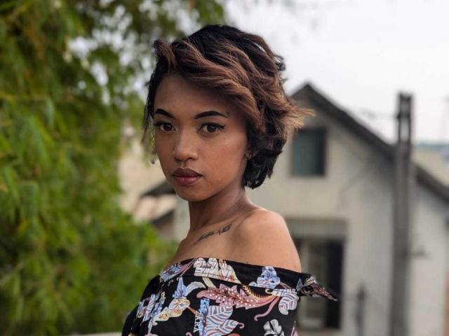 MiaLorenZ has short, wavy brown hair with blonde highlights, wearing an off-shoulder black dress with colorful patterns, showing a tattoo on her neck, looking serious.