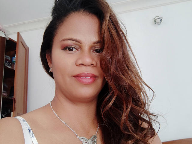 ChicetBelle with brown wavy hair, light brown skin, pink lipstick, white top, silver necklace, and small earrings, smiling slightly, showing her nose and lips.