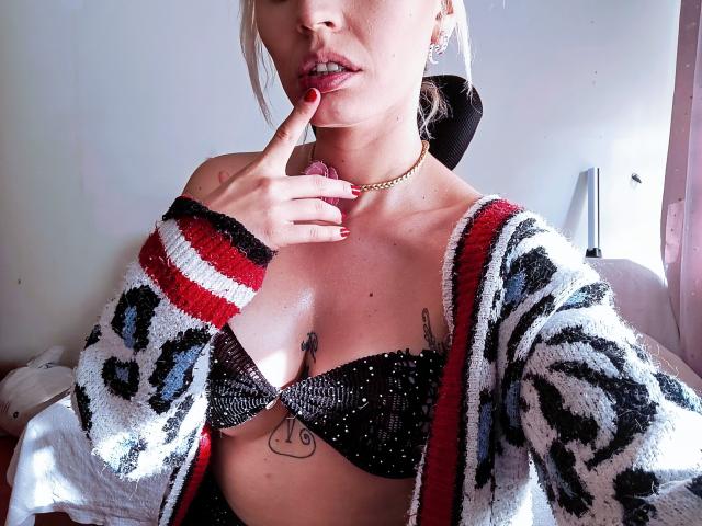 SaraleePoison with blonde hair, wearing a sparkly black bra, leopard print cardigan, gold necklace, and red nail polish, finger to her lips, showing tattoos under her tits.