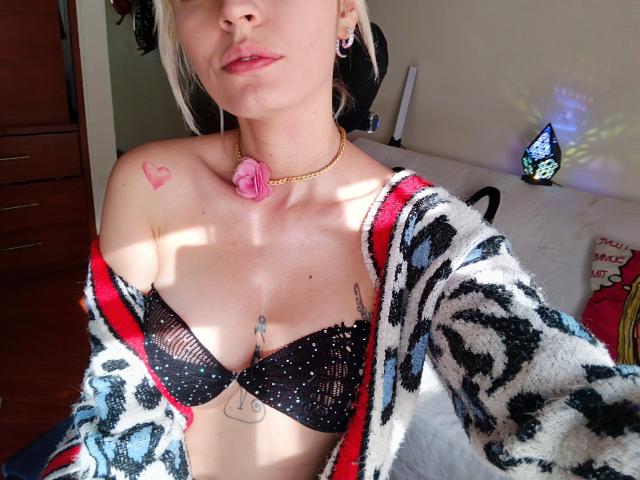 SaraleePoison with pale skin, small pink heart tattoo on right shoulder, black lace bra showing her tits, gold chain with pink rose, leopard print cardigan, and white bed.