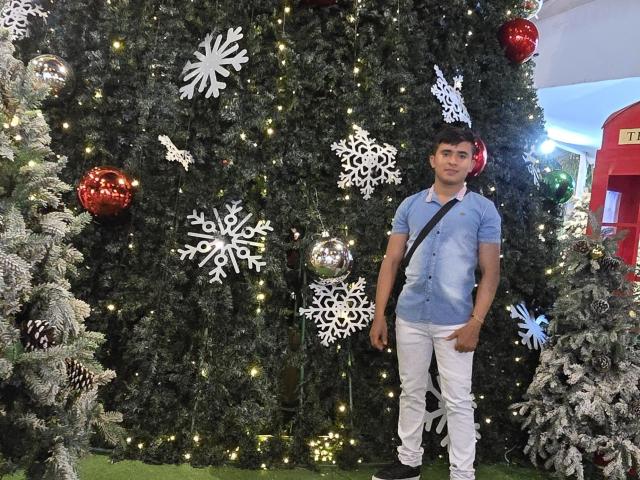 DayanWilly stands in front of a decorated Christmas tree with white snowflakes, red balls, and tiny lights, wearing a blue shirt, white pants, and a black strap.