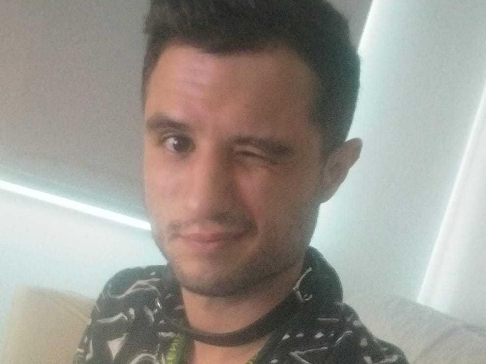 EnriqueSuave winking with one eye, short dark hair, light skin, slight beard, black patterned shirt, and a black choker, looking cute and slightly goofy.