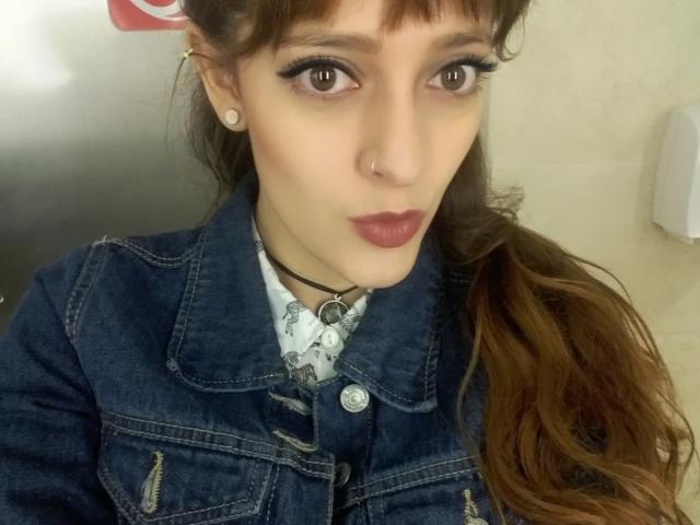 JaneRoa has brown eyes, long wavy brown hair, and is wearing a denim jacket with a black choker, pearl earrings, and dark red lipstick, showing a neutral expression.