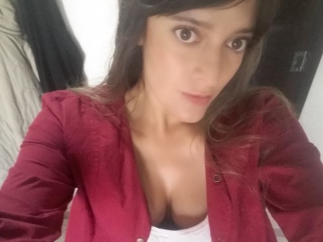 JaneRoa, met bruine ogen, draagt een rood shirt dat haar grote borsten laat zien, en een witte top. Ze heeft een lichte huid en lang bruin haar en kijkt naar de camera..
