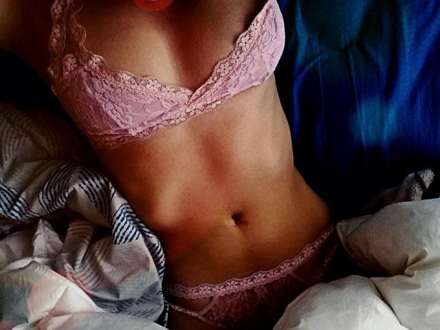 JaneRoa's slim, tan, flat-stomached body in pink lace lingerie, showing her small, perky boobs and lace-covered pussy, lying on a blue bed with a patterned blanket.