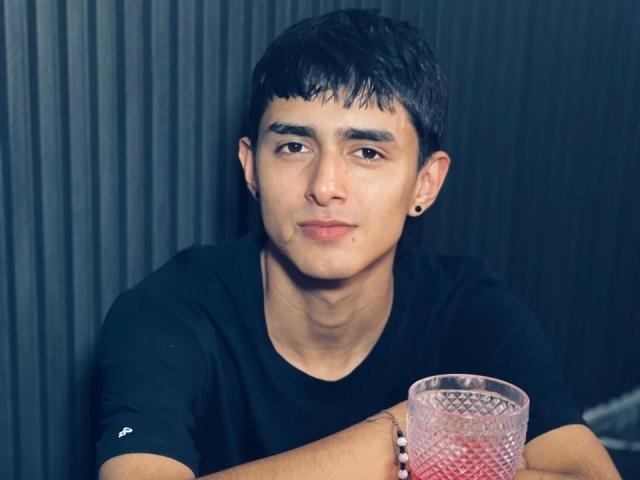 NathanMaxime, con capelli neri corti, una maglietta nera, che tiene un bicchiere rosa, con un'espressione leggermente annoiata, un piccolo orecchino nero nell'orecchio sinistro, una parete grigia a coste sullo sfondo..