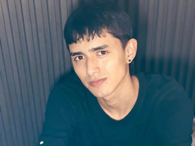 NathanMaxime has short black hair, dark eyes, and a slight smirk. He's wearing a black shirt with small white logos on the sleeves, and has a black ear stud.