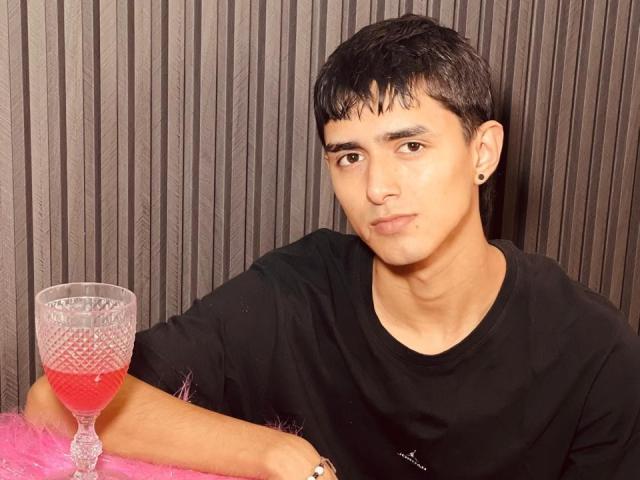 NathanMaxime with short black hair, wearing a black shirt, holding a pink glass with red liquid, sitting on pink furry surface, looking at camera with neutral expression.