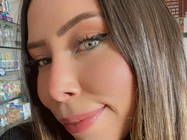 JennyLawry has green eyes, brown hair, and is wearing green eyeshadow with long eyelashes. She's smirking with pink lipstick, showing her slightly shiny face.