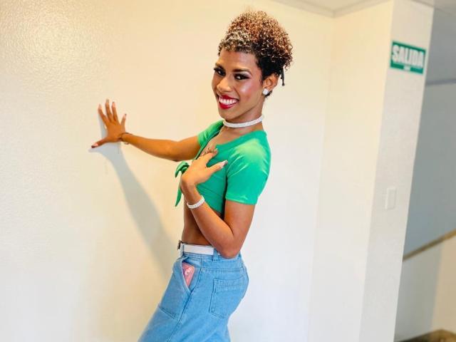 ThiaraVarannes, avec des cheveux bruns bouclés, un haut vert ajusté, un jean bleu, un collier blanc, souriante, avec du rouge à lèvres, touchant un mur blanc, la main droite sur le mur, la main gauche sur la poitrine, portant un bracelet..