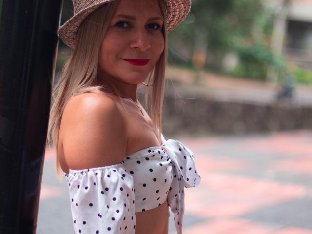 JuliethVarx with blonde hair, red lipstick, and a polka-dot off-shoulder top, wearing a straw hat, smiling at the camera, showing her tits, standing near a black pillar.