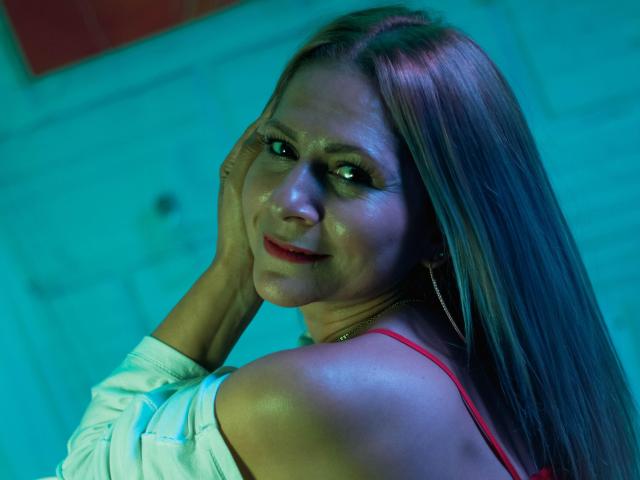 JuliethVarx, con la pelle luminosa, i lunghi capelli castani, il rossetto rosso e un sorriso malizioso, tiene il viso tra le mani, indossa una maglietta rossa e orecchini a cerchio, illuminata da una luce blu-verde..