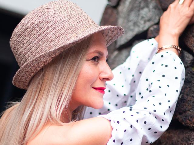 JuliethVarx with blonde hair, wearing a pink woven hat and white polka dot shirt, touching a rock, red lipstick, smiling, gold watch on wrist.