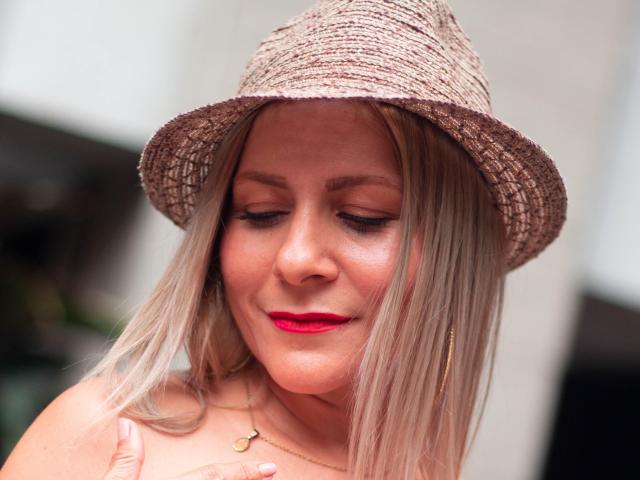JuliethVarx with blonde hair, wearing a straw hat, red lipstick, and a gold necklace, covering her bare boobs with her hand, smiling softly, looking downward.