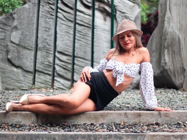 JuliethVarx indossa una maglietta a maniche corte con pois, una gonna nera, un cappello a secchiello beige e tacchi a spillo, è sdraiata sulla ghiaia, mostrando il seno, i glutei e il rossetto rosso, con un sorriso rilassato..