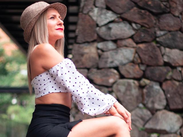 JuliethVarx, con i capelli biondi, il rossetto rosso e un cappello di paglia, indossa una maglietta a pois con una spalla scoperta e una gonna nera, seduta di fronte a un muro di pietra..