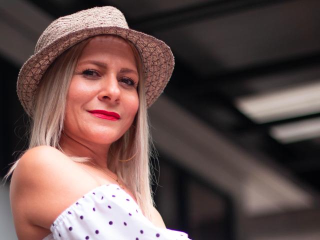 JuliethVarx, con i capelli biondi, indossa un cappello di paglia e rossetto rosso, sorridendo con una maglietta bianca a spalle scoperte con pois viola, che mostra leggermente il seno..