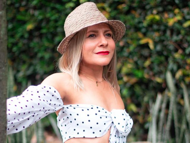 JuliethVarx, con i capelli biondi, il rossetto rosso e un cappello di paglia, indossa una maglietta bianca a pois con una spalla scoperta, che mette in mostra il suo seno piccolo e la sua figura snella, ed è in piedi all'aperto..