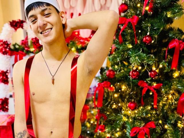 LucianMael, shirtless, wearing red suspenders, white headband, smiling, winking, hand behind head, small tattoo, necklace, Christmas tree with red ornaments and lights in background.