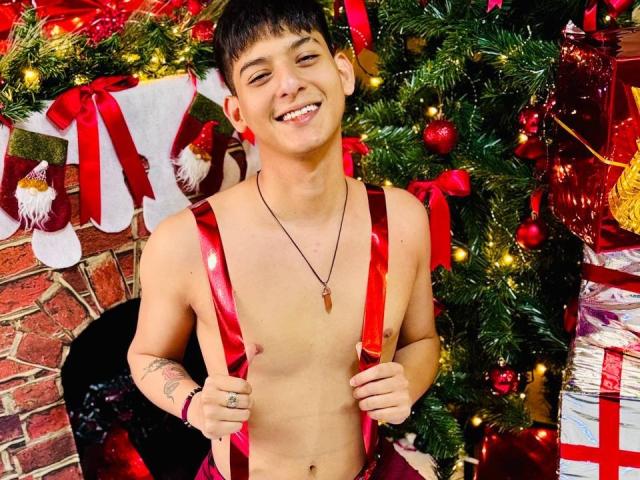 LucianMael, shirtless with red suspenders, smiling at camera, Christmas tree with red ornaments and lights in background, necklace with small cross, tattoo on right wrist, short dark hair.