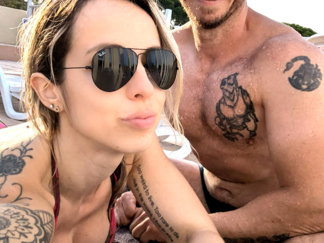 Annic with wet blonde hair, aviator sunglasses, and a red bikini top, next to a tattooed, shirtless, muscular guy with a beard and black briefs. Sunlit, outdoor setting.