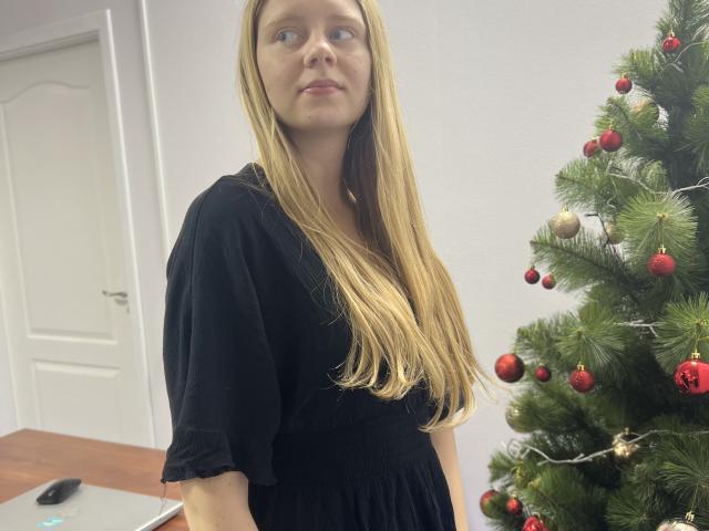 Laliina, con lunghi capelli biondi, indossa un vestito nero ed è in piedi accanto a un albero di Natale decorato con ornamenti rossi e dorati. Le sue natiche sono parzialmente visibili..