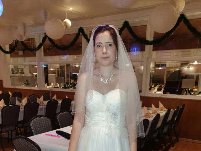 Spielzeuglena-hot stands in a room, wearing a white wedding dress with a veil, strapless, and a silver necklace. Her tits are small, and she has a cute, fair-skinned face.