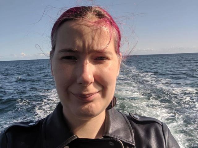 Spielzeuglena-hot with pink-dyed hair, leather jacket, and a goofy smile, standing in choppy water, sunlit face, and slightly messy hair.