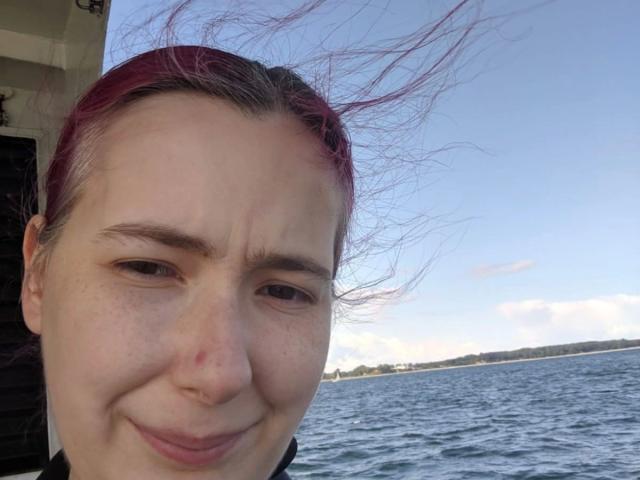 Spielzeuglena-hot's face with pink-dyed hair, wind blowing, slightly annoyed smile, small red mark on cheek, blue sky, and water in background. She's wearing a black shirt.
