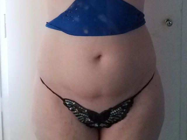 Spielzeuglena-hot's flat stomach, pale skin, small black lace thong with butterfly pattern, and blue top covering her tits, standing in front of a white door.