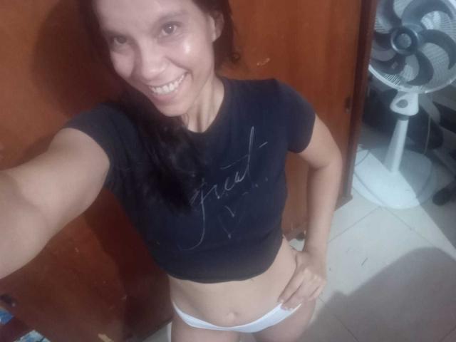 EstrellaFandy with brown hair, smiling, wearing a black shirt and white panties, standing with one hand on her hip, a fan in the background, and a wooden door.