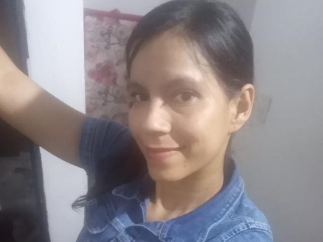 EstrellaFandy with dark hair, brown eyes, and a subtle smile, wearing a blue denim shirt, has a small nose and a cute face, her arm raised slightly.