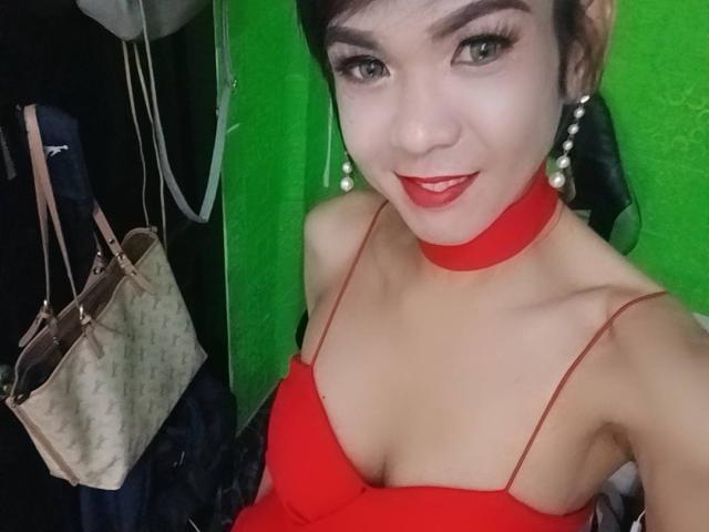 WifeMaterial with red dress, red choker, and pearl earrings, smiling at the camera, showing her tits, wearing red lipstick, and dark hair in a side ponytail.