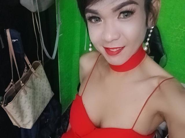 WifeMaterial with red dress, choker, and pearl earrings, smiling at camera, showing small tits, red lipstick, and black hair, green background, Louis Vuitton bag on left.