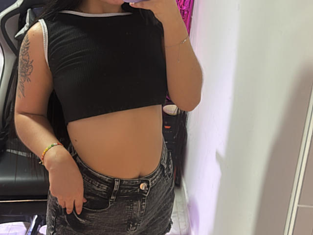 SarahGood with a black crop top, showing her flat midriff and tattooed arm, wearing gray denim shorts, holding a phone, and a colorful bracelet.