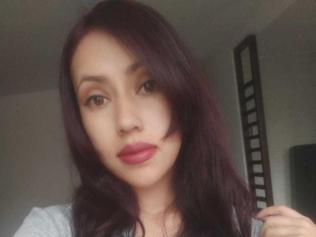 MariaMendozaI has dark purple hair, brown eyes, and red lipstick, with a serious expression, looking at the camera, wearing a grey top.