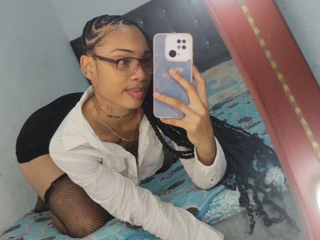 ElaHott, con trenzas largas, gafas, lengua afuera, vistiendo una camisa blanca, pantalones cortos negros, medias de red, tomando un selfie, mostrando su piel bronceada, un tatuaje pequeño en el cuello y una cara linda..