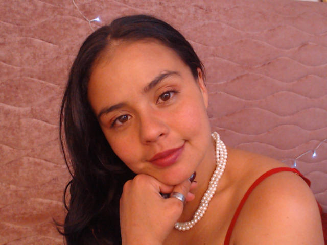 ArielleWilde with dark hair, brown eyes, and a pearl necklace, resting her chin on her hand, wearing a red strap, smiling gently. Her tits are partially visible.