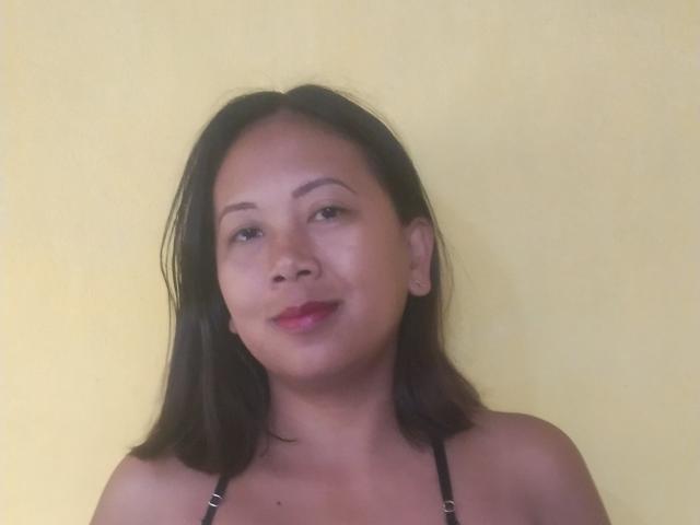 SarahFontaineX has medium brown skin, black shoulder-length hair, and is smiling slightly with red lipstick. She's wearing a black strap top. Her tits are small.