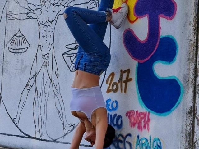 JuneLaFrenchie hangs upside down on a graffiti-covered wall, wearing tight blue jeans and white panties, exposing a toned midriff. Her legs are spread, showcasing a sexually provocative pose.