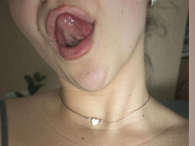 La bocca di SugarrX è aperta, con la lingua fuori, mostrando labbra rosa lucide e leggermente umide, e un collo parzialmente visibile con una collana a forma di cuore. La sua pelle è pallida, con piccole lentiggini..