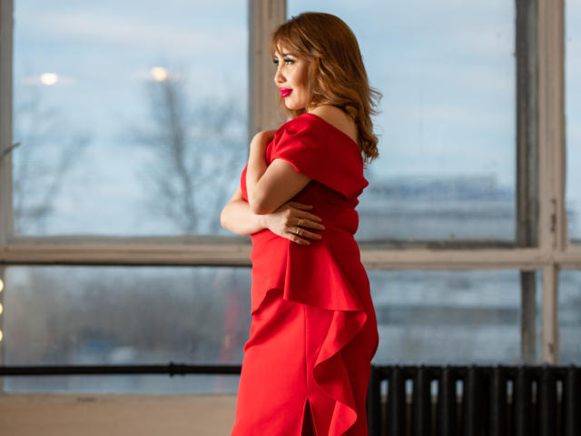 LiliNi stands in a red dress, arms crossed, showing her tits, with red lipstick and wavy brown hair, in front of a large window.