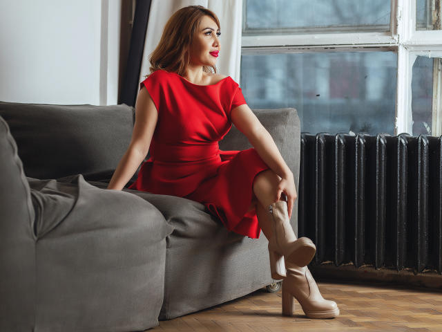 LiliNi sits on a gray couch, wearing a red dress and beige boots, with red lipstick, looking to the side. She's touching her knee, showing off her tits.