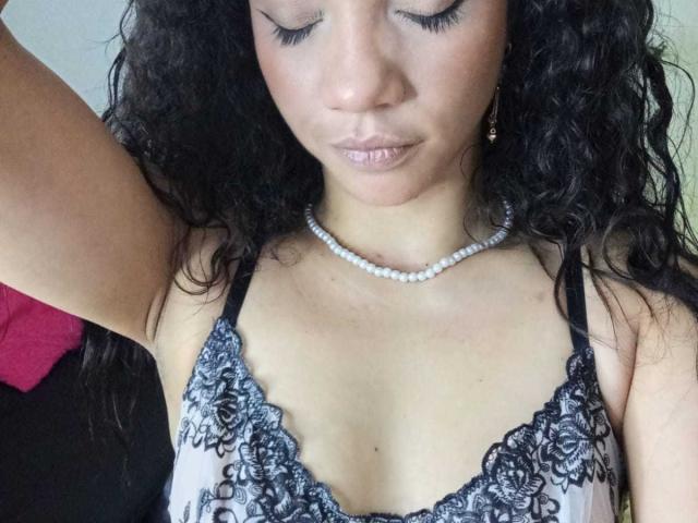 AndromedaZareth has curly black hair, wearing a lace-trimmed top, pearl necklace, and eyeliner. Her eyes are closed, and her tits are small with a subtle cleavage.