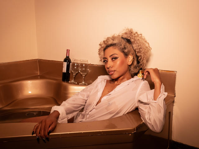 LilithWalker with curly blonde hair, tan skin, wearing a white shirt, in a gold bathtub, holding a wine bottle and glasses, looking seductively at the camera.