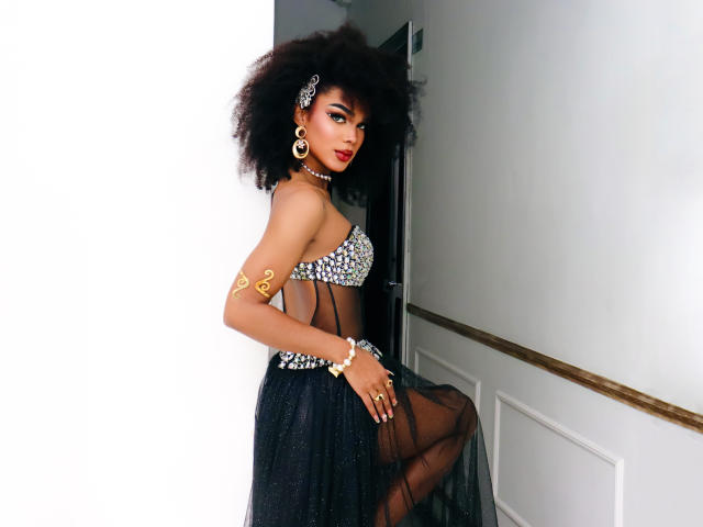 NaomiSteward with big afro, wearing a sparkly bra, sheer black skirt, gold arm tattoos, and jewelry, leaning against a white wall, showing her small tits and red lipstick.
