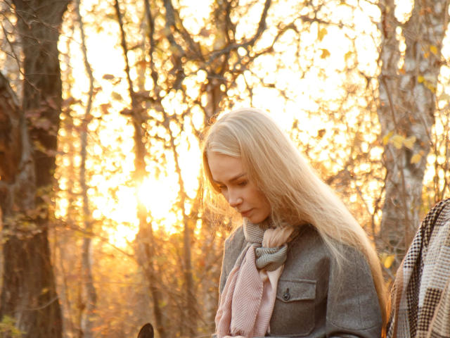 BlondiAngel: una ragazza attraente con lunghi capelli biondi lisci, che indossa un cappotto grigio e una sciarpa rosa, con lo sguardo rivolto verso il basso, la luce del sole filtra tra gli alberi autunnali, una luce dorata calda, un'espressione dolce e serena..