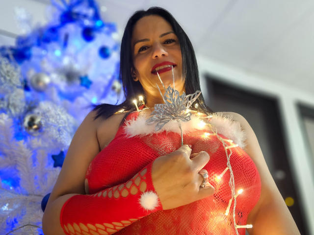 LizaRussell with black hair, red fishnet dress, and Christmas lights, holding them against her large tits, standing in front of a blue Christmas tree. She's smiling with red lipstick.