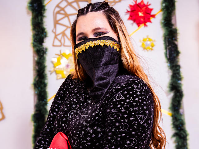 SalmaAzaman with long, wavy brown hair, black mask with yellow trim, black patterned shirt, covering her mouth, holding red object, in front of festive, colorful background.